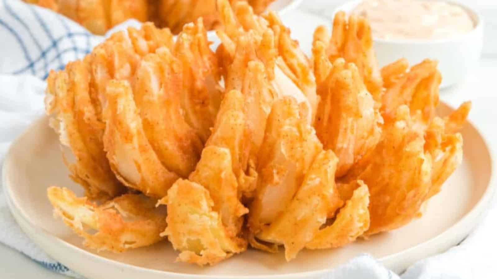 A golden, crispy blooming onion sits on a plate with dipping sauce in the background.