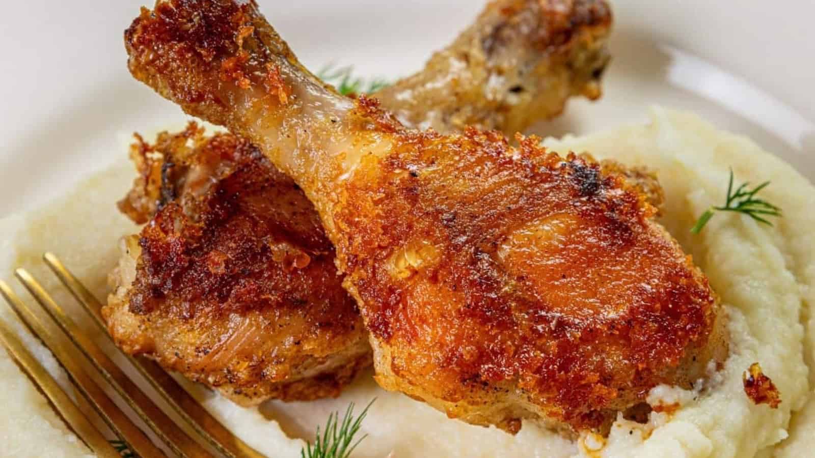 Two crispy fried chicken drumsticks on a bed of creamy mashed potatoes, garnished with dill. A fork is placed beside the dish. The chicken is golden brown and the potatoes have a smooth texture.