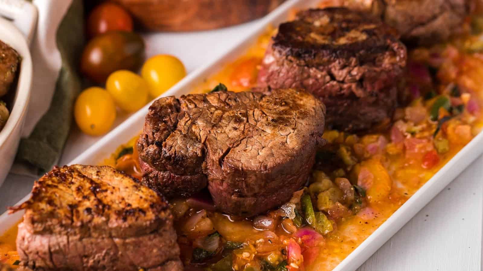 Three seared filet mignon steaks served on a platter over a chunky tomato and vegetable sauce.