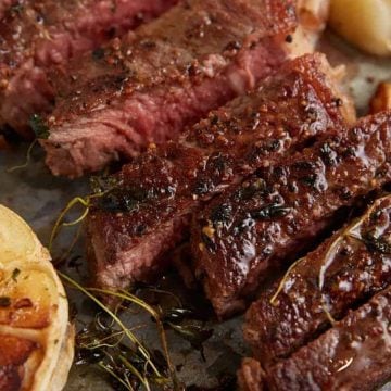 Sliced medium-rare steak with herbs, garlic, and seasoning on a rustic surface.