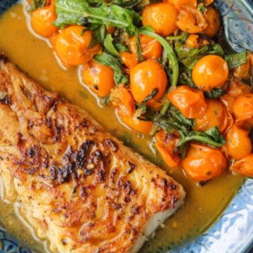 Grilled fish fillet with sauce, served with sautéed cherry tomatoes and greens on a blue plate.
