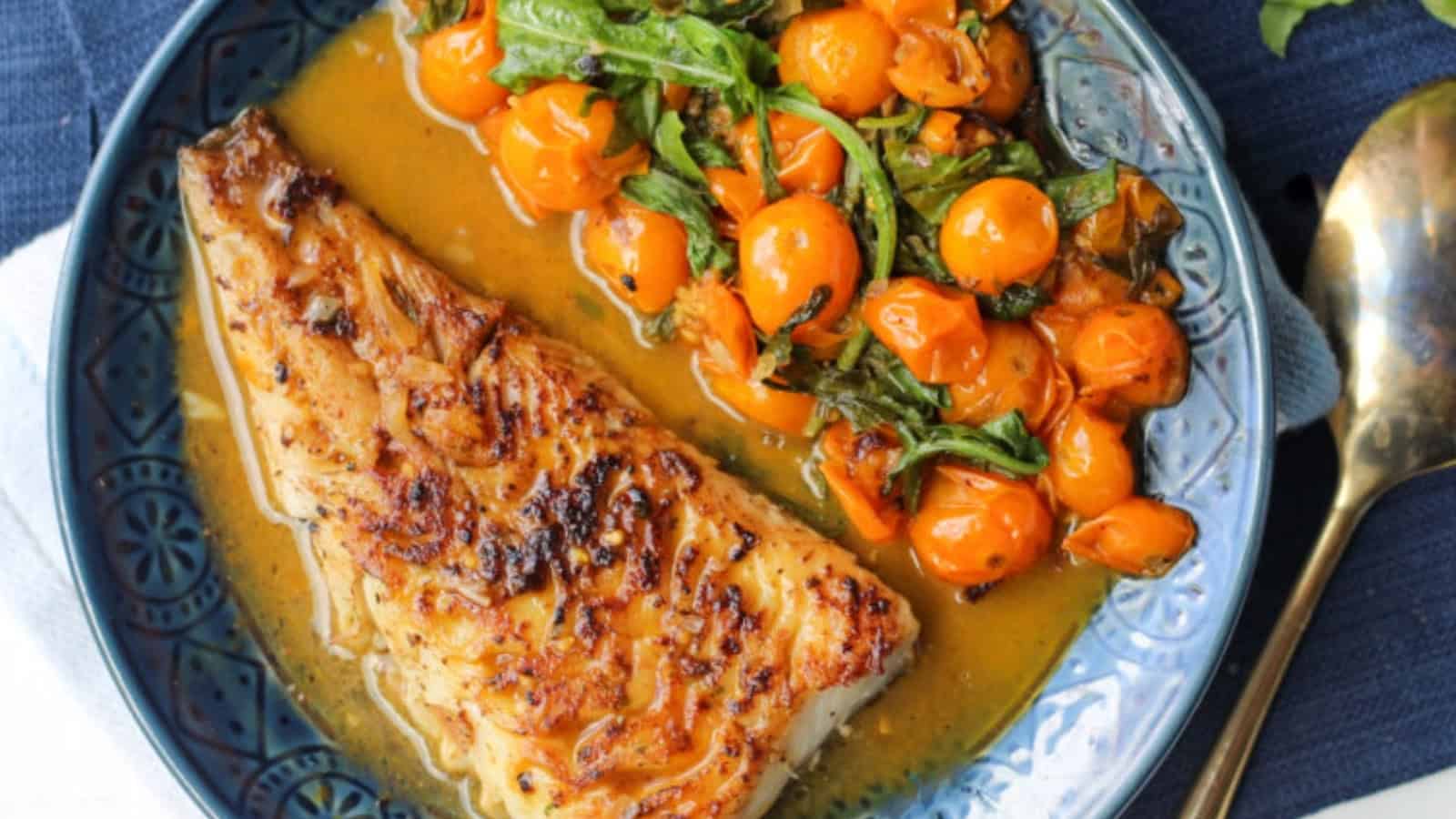 Grilled fish fillet with sauce, served with saut&eacute;ed cherry tomatoes and greens on a blue plate.