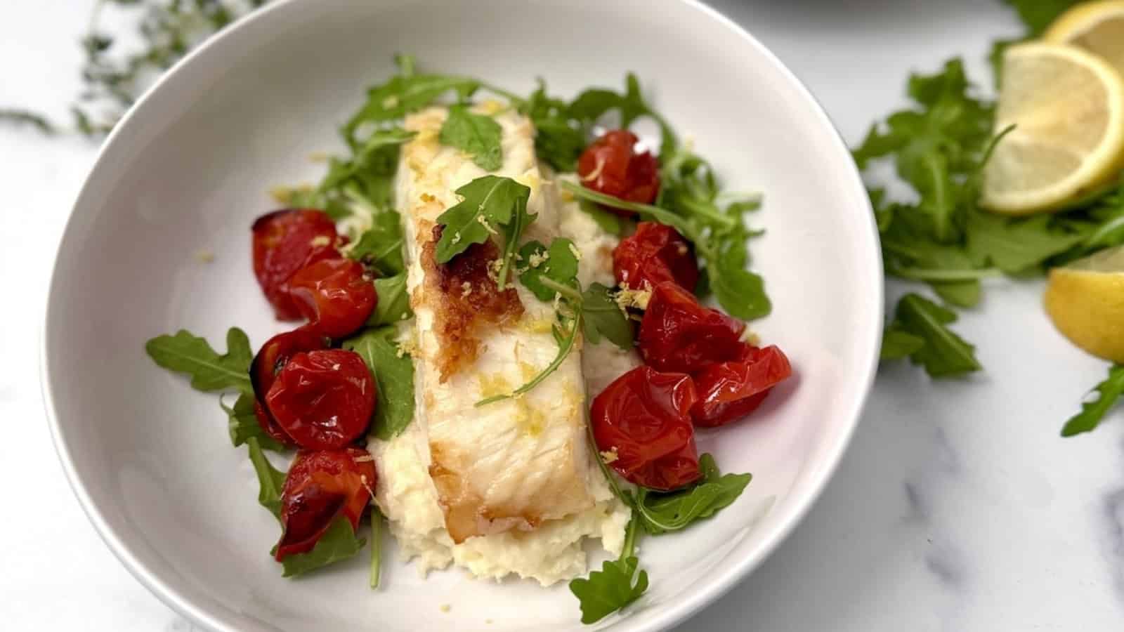 White fish fillet on mashed potatoes with roasted cherry tomatoes and arugula, garnished with lemon slices.