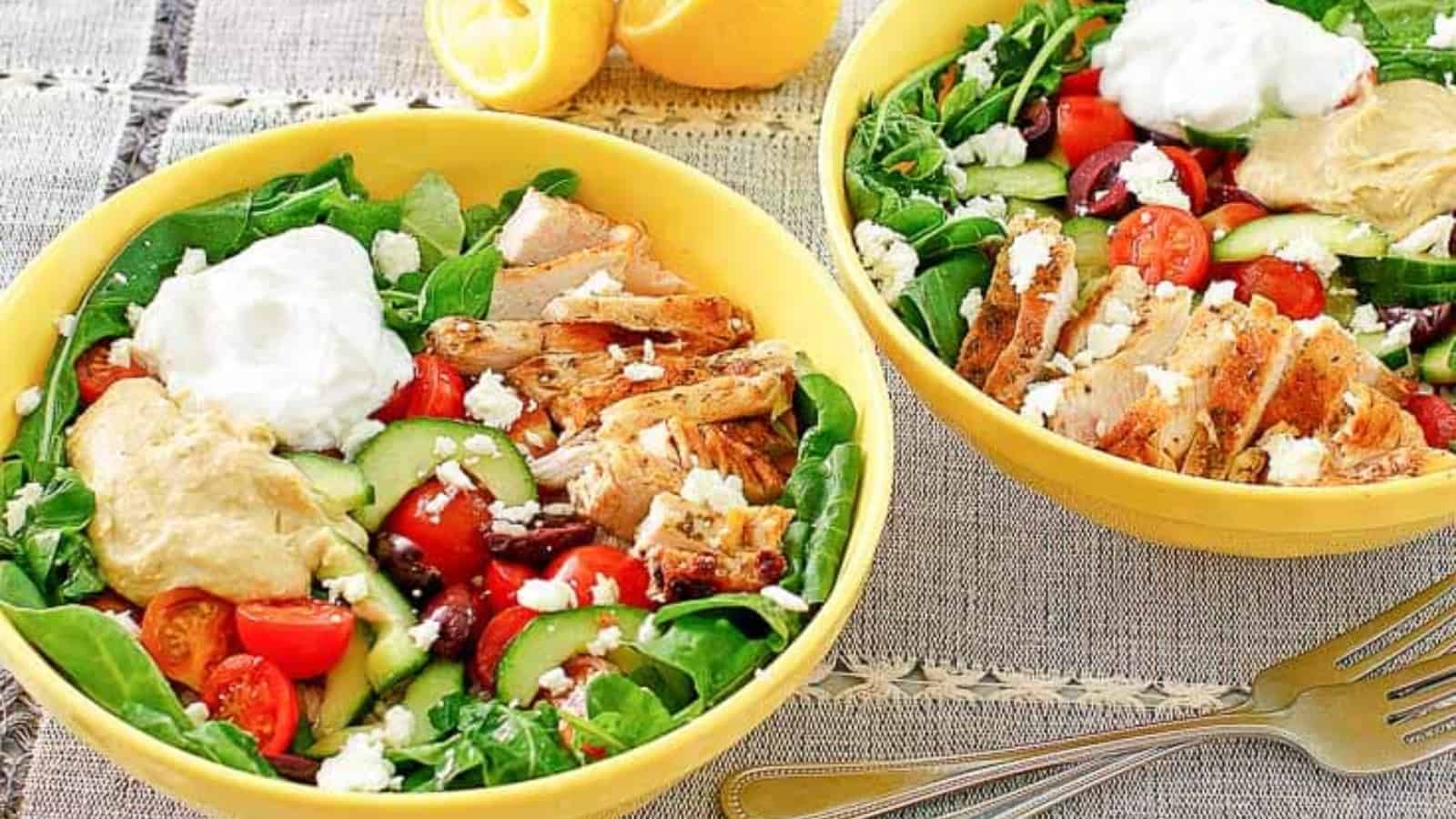 Two yellow bowls with Greek salad, grilled chicken, hummus, tzatziki, tomatoes, cucumber, and feta cheese.