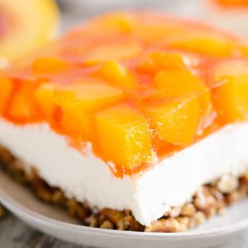A dessert bar with a crumbly base, creamy middle layer, and peach chunks in a gelatin topping on a plate.