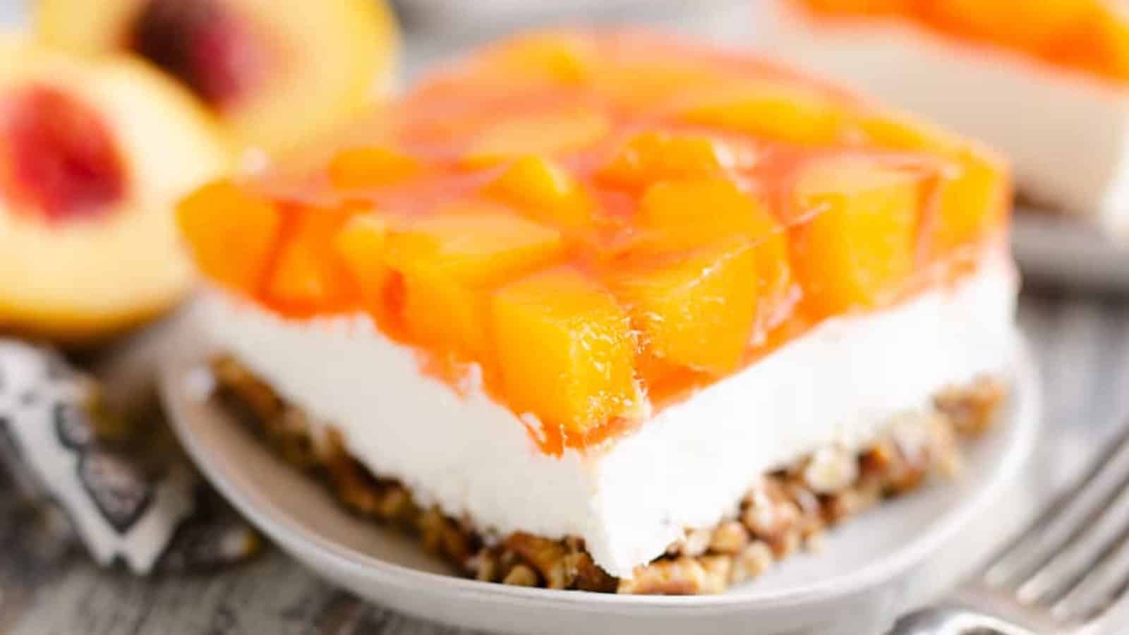 A dessert bar with a crumbly base, creamy middle layer, and peach chunks in a gelatin topping on a plate.