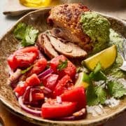 Roast chicken with green sauce, tomato and red onion salad, lime wedge, and cilantro on a ceramic plate.