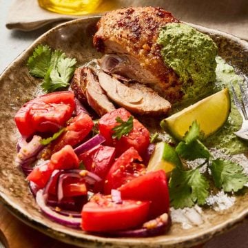 Roast chicken with green sauce, tomato and red onion salad, lime wedge, and cilantro on a ceramic plate.