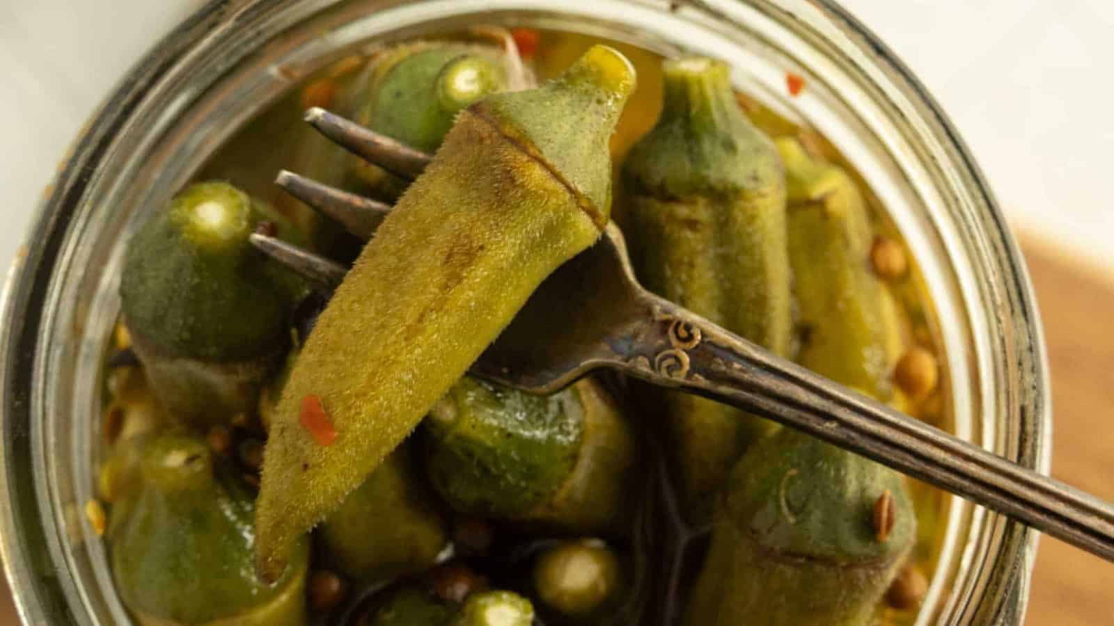 A fork holding a piece of pickled okra above a jar filled with more pickled okra.
