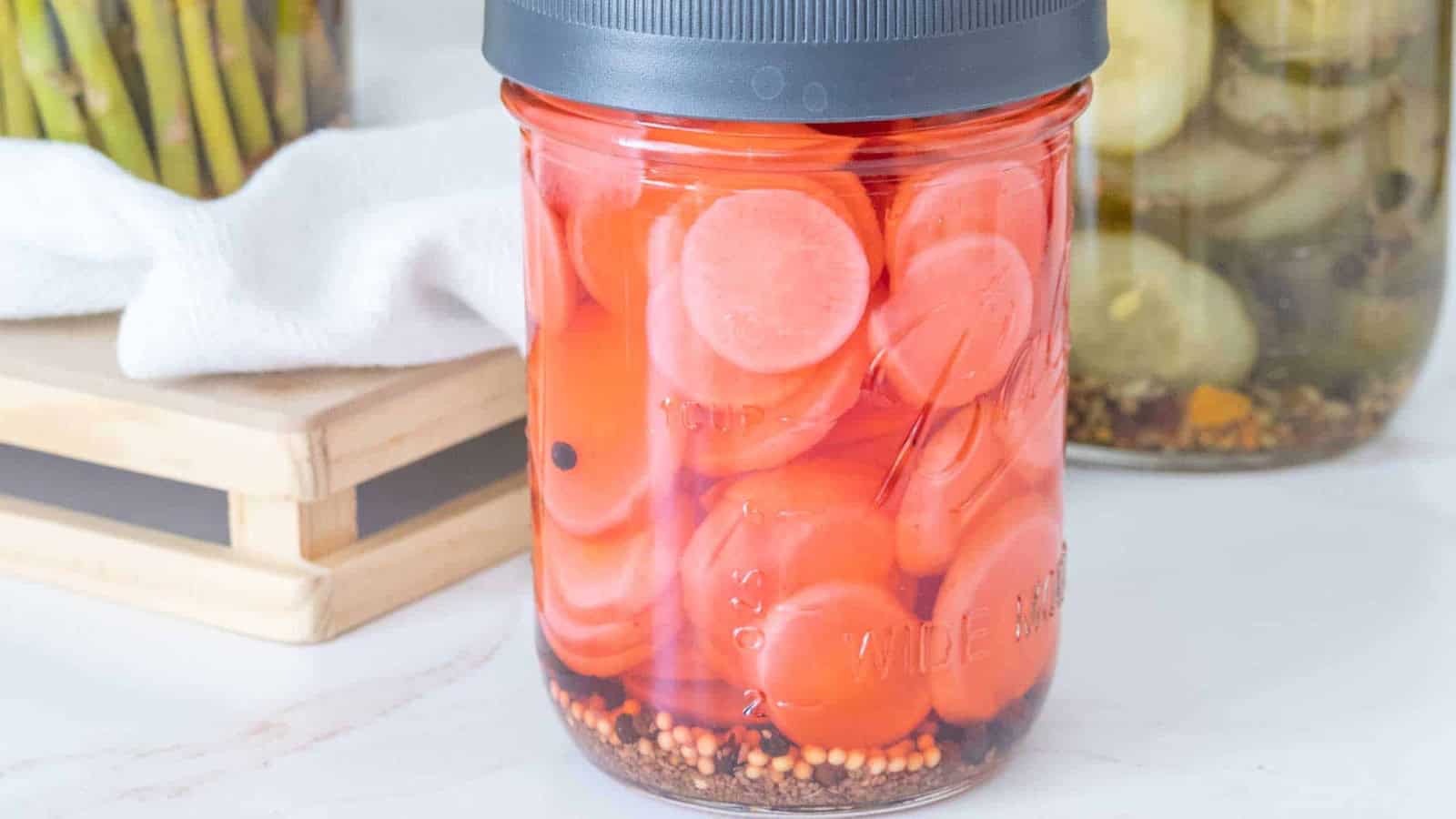 A jar of pickled carrots with spices at the bottom, sitting on a white surface near other jars.