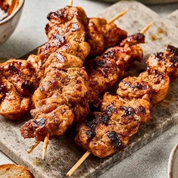 Grilled chicken skewers with spices on a cutting board, with sauce and bread nearby.