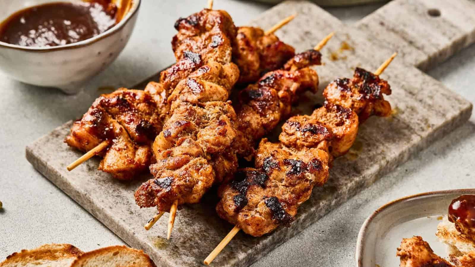 Grilled chicken skewers with spices on a cutting board, with sauce and bread nearby.