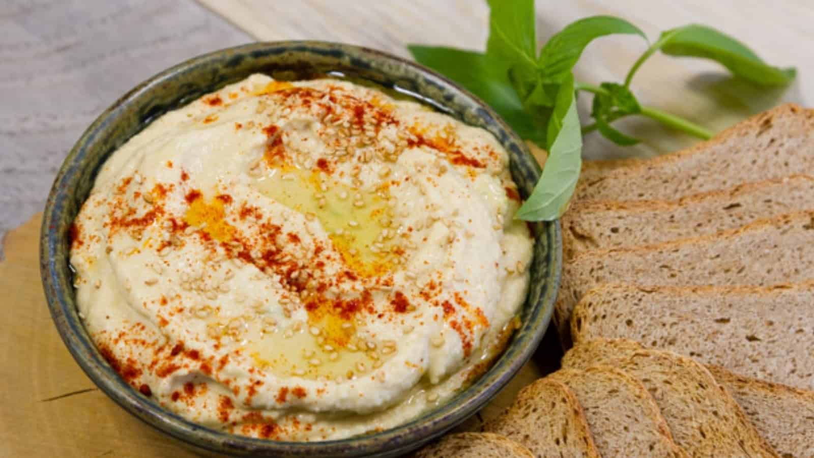 A bowl of hummus with paprika and oil, garnished with sesame seeds, beside sliced brown bread and basil.