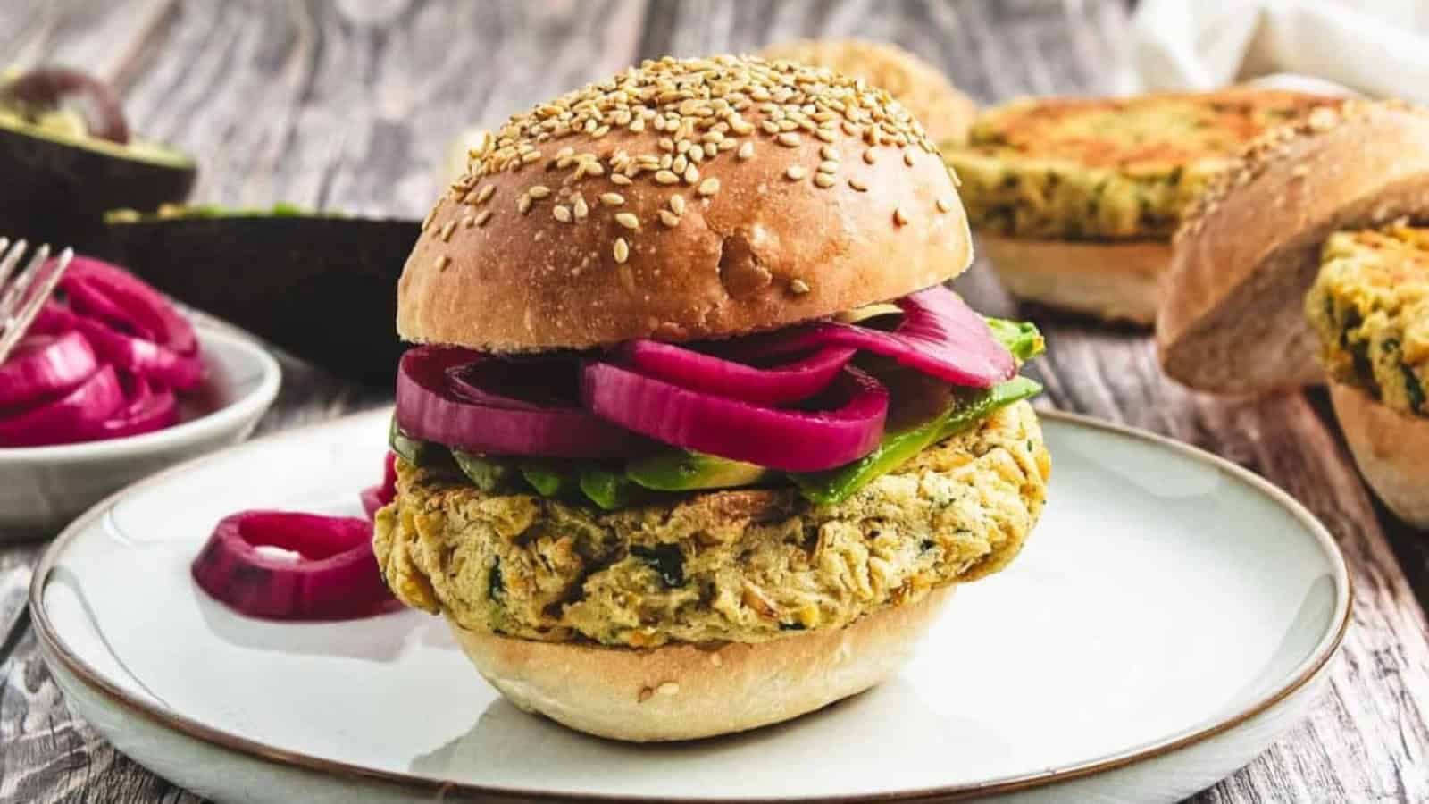 A veggie burger with pickled onions and avocado on a sesame bun sits on a white plate.