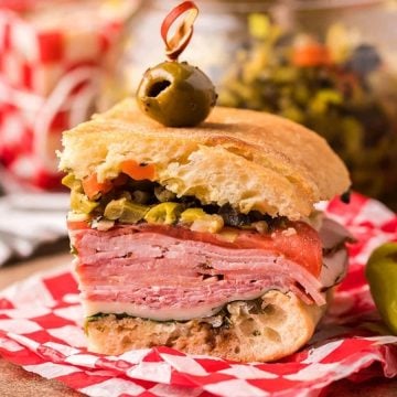 A stacked Italian sandwich with meats, cheese, and olive salad, topped with an olive, on red checkered paper.