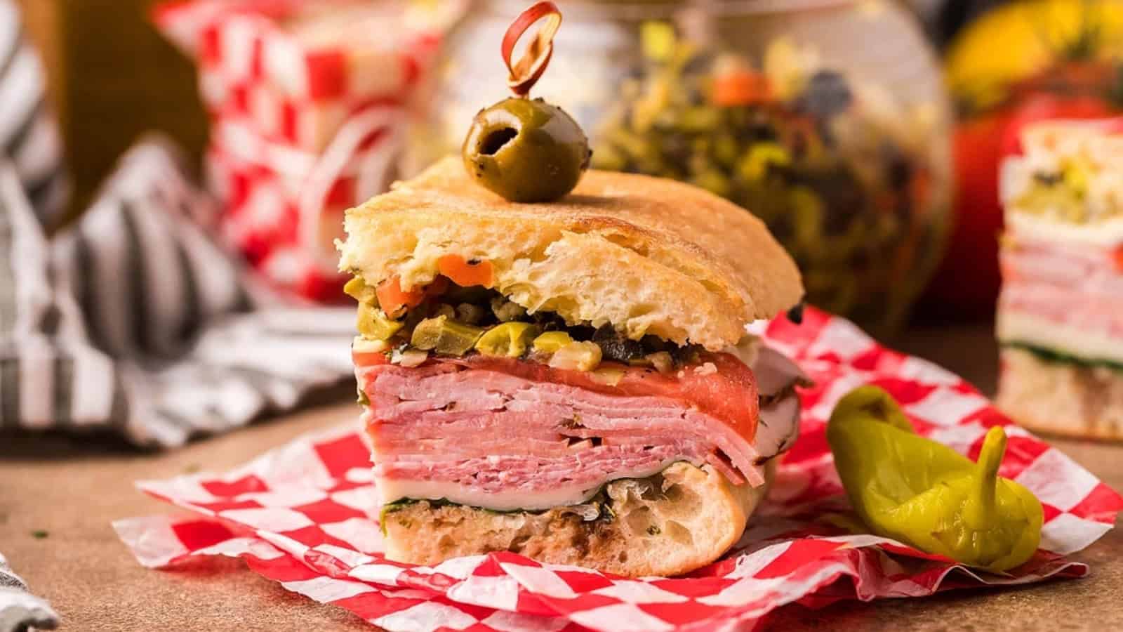 A stacked Italian sandwich with meats, cheese, and olive salad, topped with an olive, on red checkered paper.