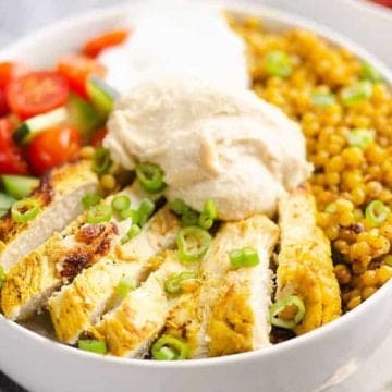 A bowl with sliced chicken, sliced green onions, hummus, tomatoes, cucumber, lentils, and a lemon wedge.