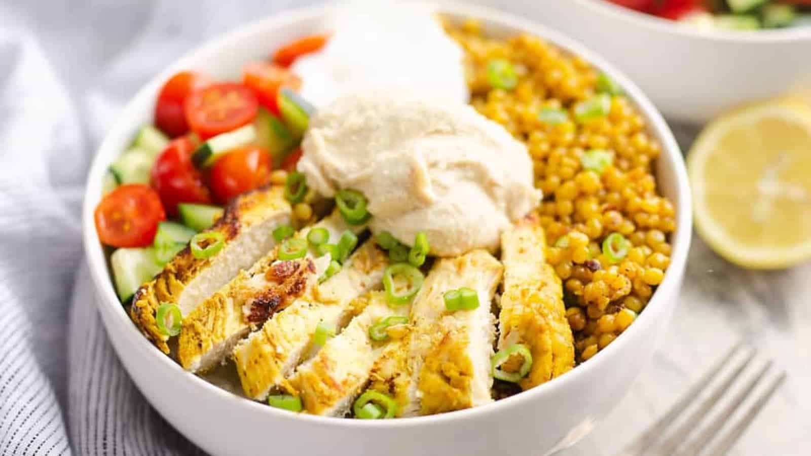 A bowl with sliced chicken, sliced green onions, hummus, tomatoes, cucumber, lentils, and a lemon wedge.