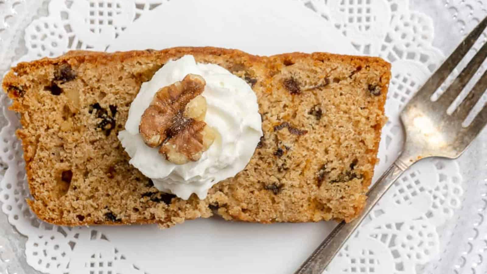 Slice of walnut cake topped with whipped cream and a walnut, on a plate with a fork and lace doily.