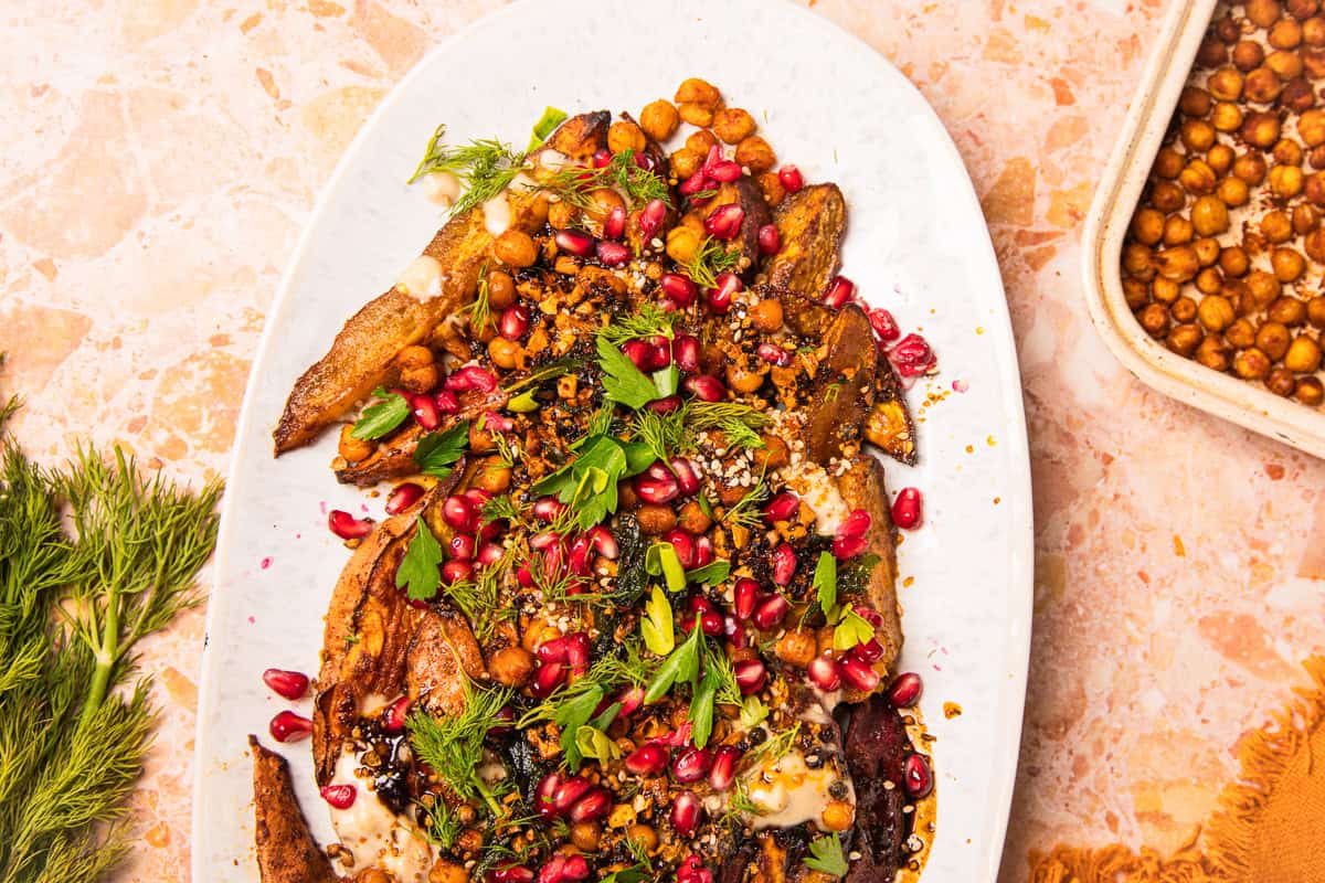 Roasted eggplant topped with chickpeas, pomegranate seeds, herbs, and sauce on a white plate, served alongside roasted sweet potatoes with tahini for a flavorful, vibrant presentation.