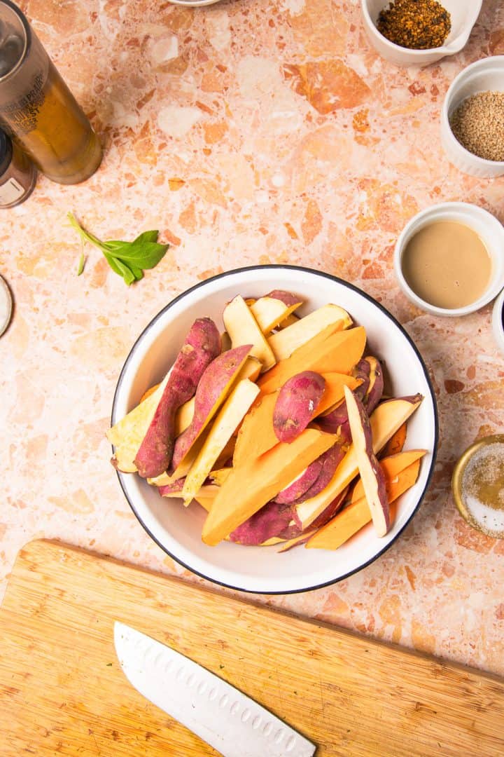 Bowl of sliced sweet potatoes on a counter with spices, sauce, and a knife on a wooden board nearby, perfect for preparing roasted sweet potatoes with tahini.