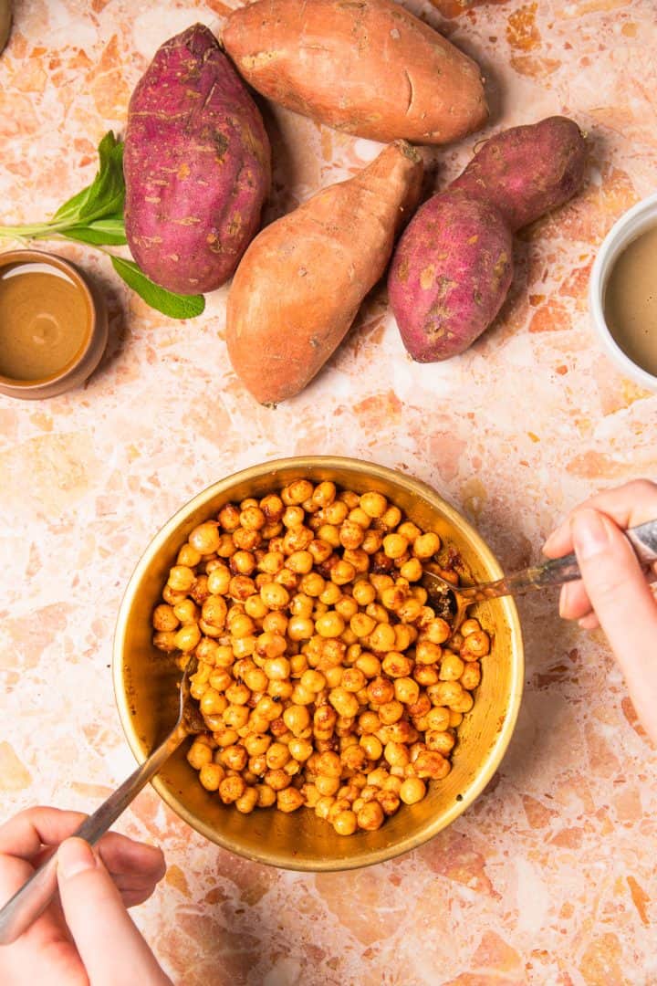 A bowl of roasted chickpeas is mixed by hand, with roasted sweet potatoes with tahini and sauce bowls nearby on a countertop.