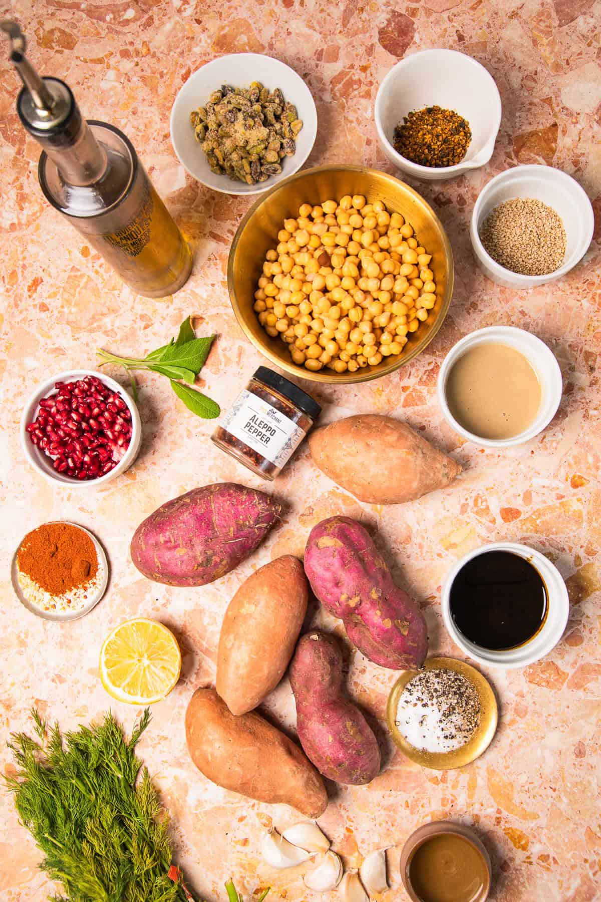 Sweet potatoes, chickpeas, spices, and various bowls of seasonings on a marble countertop, ready to be transformed into flavorful roasted sweet potatoes with tahini.