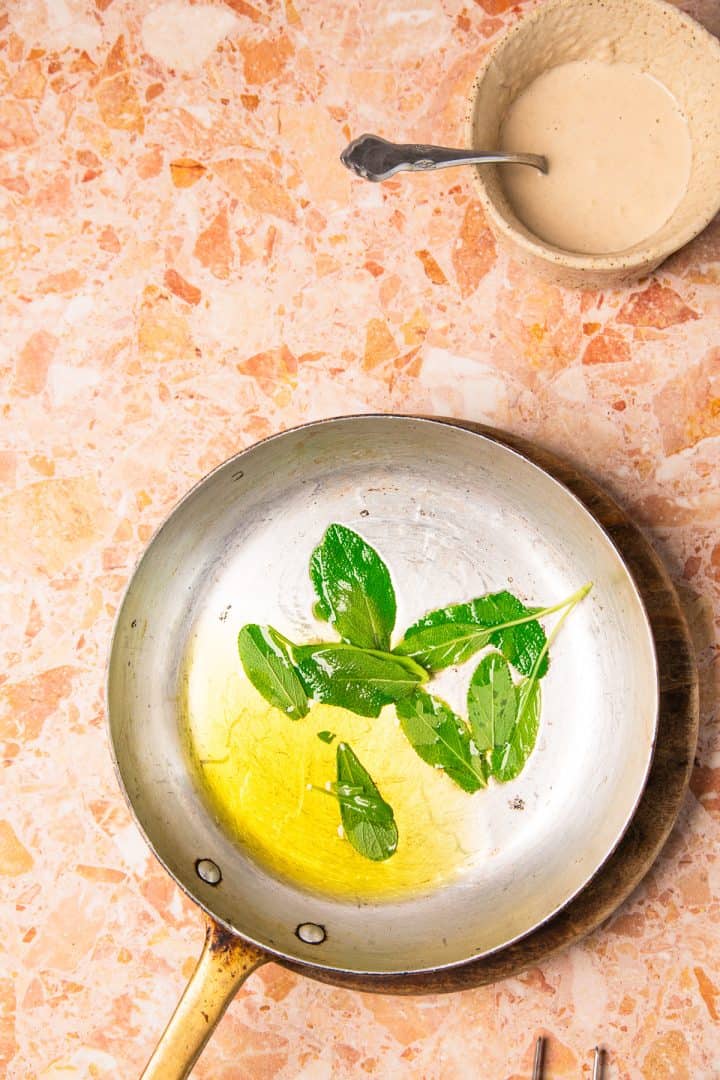 A frying pan with oil and fresh herbs beside a bowl of sauce on a pink marble countertop, ready for preparing roasted sweet potatoes with tahini.