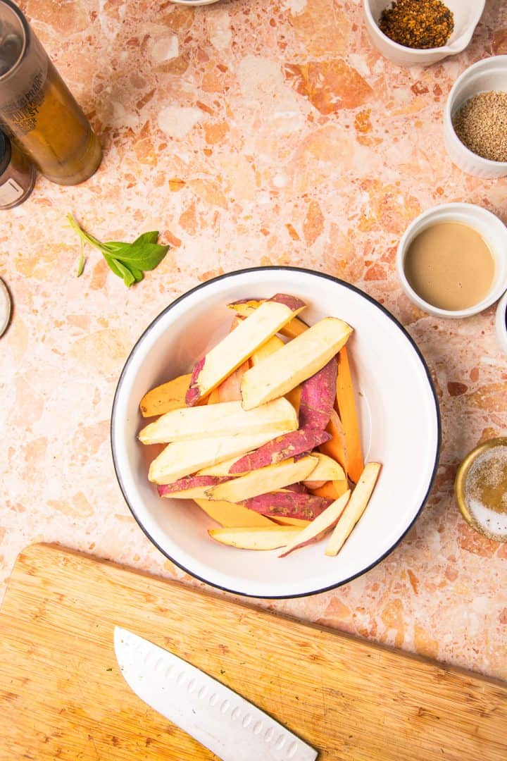 Bowl of raw sweet potato fries on a counter with seasonings, a chopping board, and a knife nearby-perfectly prepped for making roasted sweet potatoes with tahini.