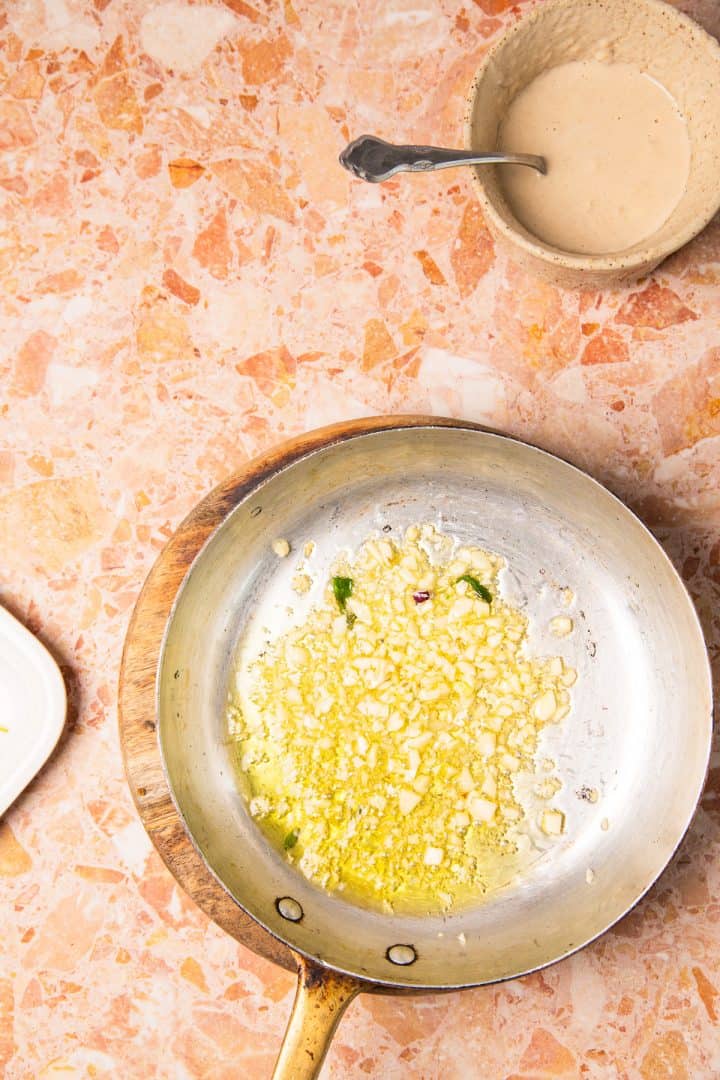 A frying pan with chopped garlic sizzling in oil beside a bowl of sauce with a spoon on a stone countertop, ready to complement roasted sweet potatoes with tahini.