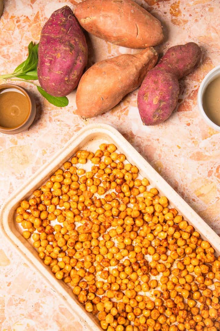 Tray of roasted chickpeas, roasted sweet potatoes with tahini, and two small bowls of sauce on a marble surface.