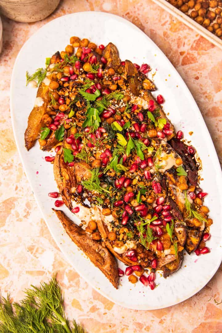 Roasted sweet potato wedges with tahini, chickpeas, pomegranate seeds, and fresh herbs are beautifully arranged on a white platter and drizzled with savory sauce.