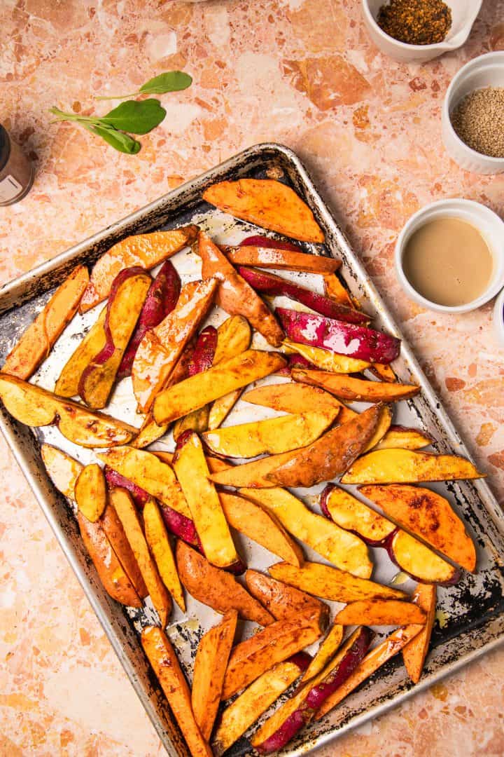 Baked sweet potato wedges on a baking sheet, surrounded by small bowls of spices and sauce-these roasted sweet potatoes with tahini are perfect for dipping and sharing.