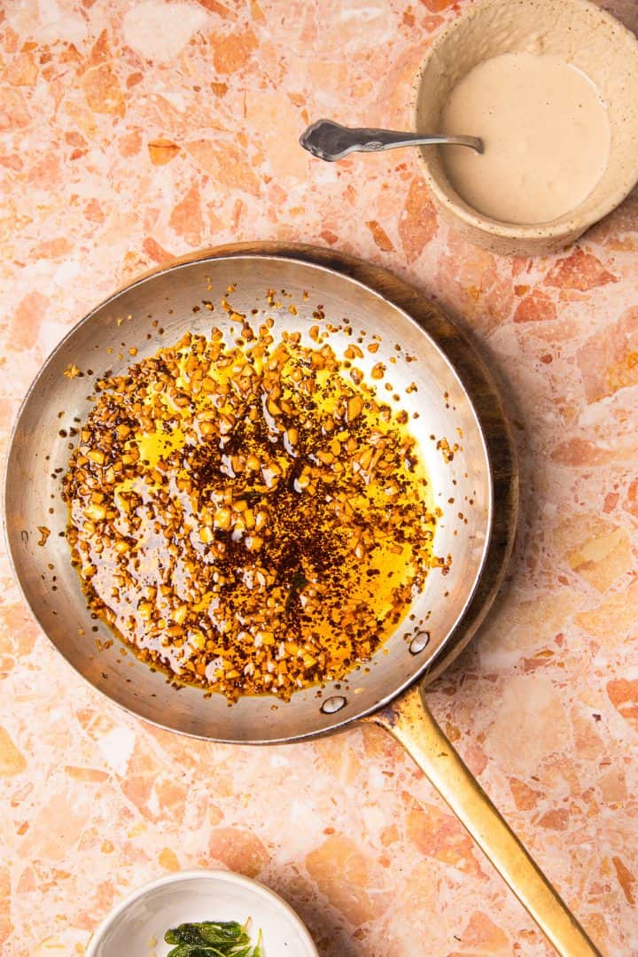 A skillet with sizzling garlic in oil sits next to a bowl of sauce and a small dish of roasted sweet potatoes with tahini on a marble countertop.