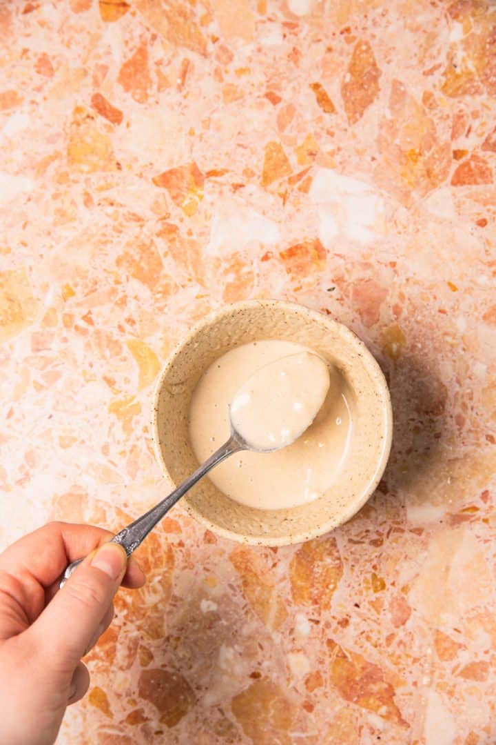 A hand holds a spoon over a small bowl of creamy sauce on a marbled orange and white countertop, ready to drizzle it atop roasted sweet potatoes with tahini.