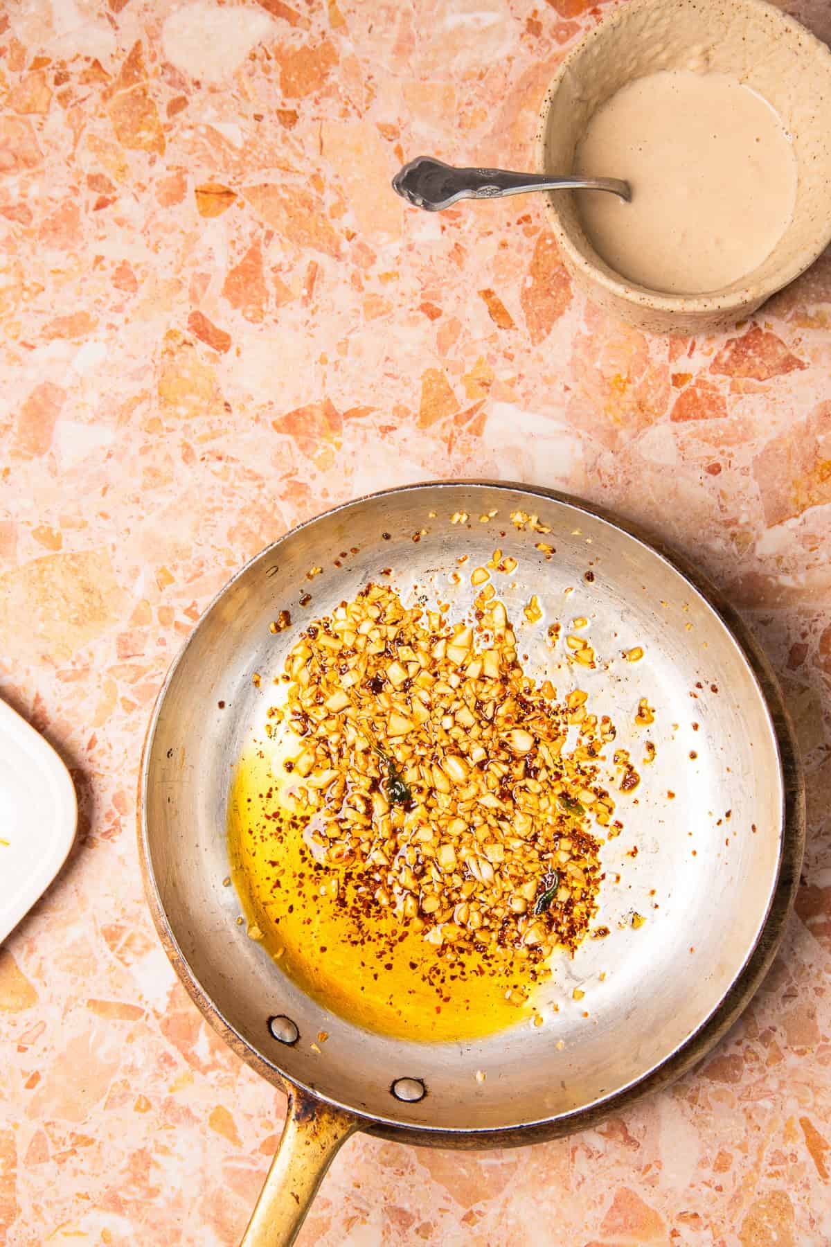 A pan with sizzling chopped garlic and oil on a pink countertop, perfect for preparing roasted sweet potatoes with tahini, with sauce in a bowl nearby.