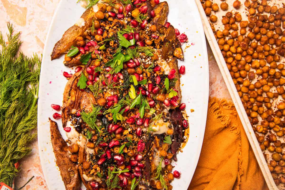 Roasted sweet potatoes with tahini, chickpeas, pomegranate seeds, fresh herbs, and spices are artfully arranged on a white platter.