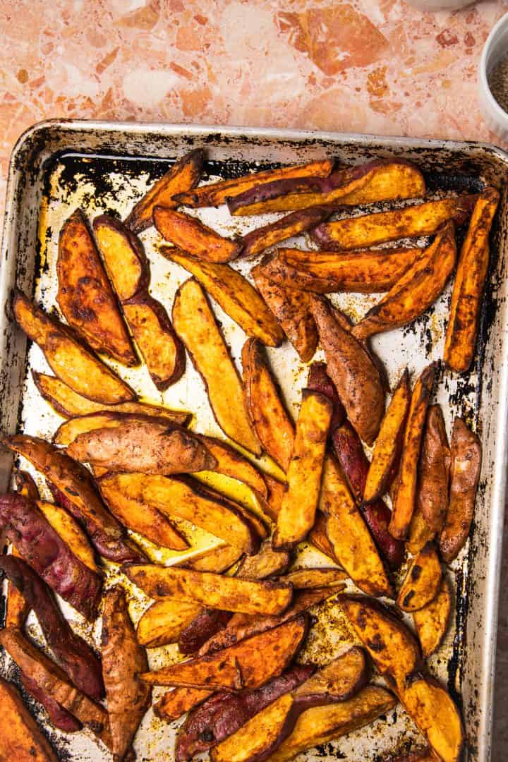 Baked potato wedges on a metal baking sheet, seasoned and golden brown, sit alongside roasted sweet potatoes with tahini on a marble countertop.