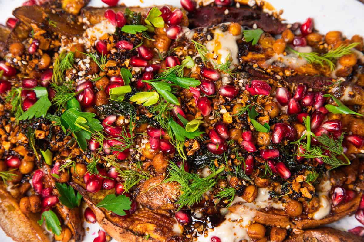 Roasted eggplant topped with chickpeas, pomegranate seeds, herbs, and spices on a white plate, served alongside roasted sweet potatoes with tahini for a deliciously vibrant meal.