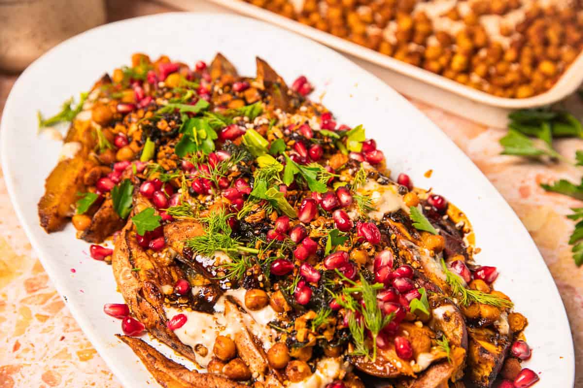 Roasted sweet potatoes with tahini are topped with chickpeas, fresh herbs, pomegranate seeds, and a creamy drizzle on a platter.