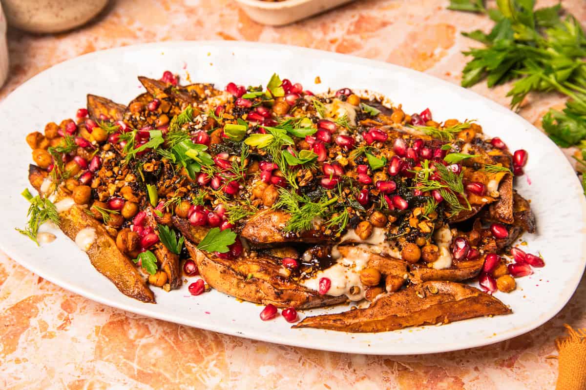 Roasted potato wedges topped with chickpeas, pomegranate seeds, fresh herbs, and a creamy tahini sauce on a white plate, reminiscent of roasted sweet potatoes with tahini.