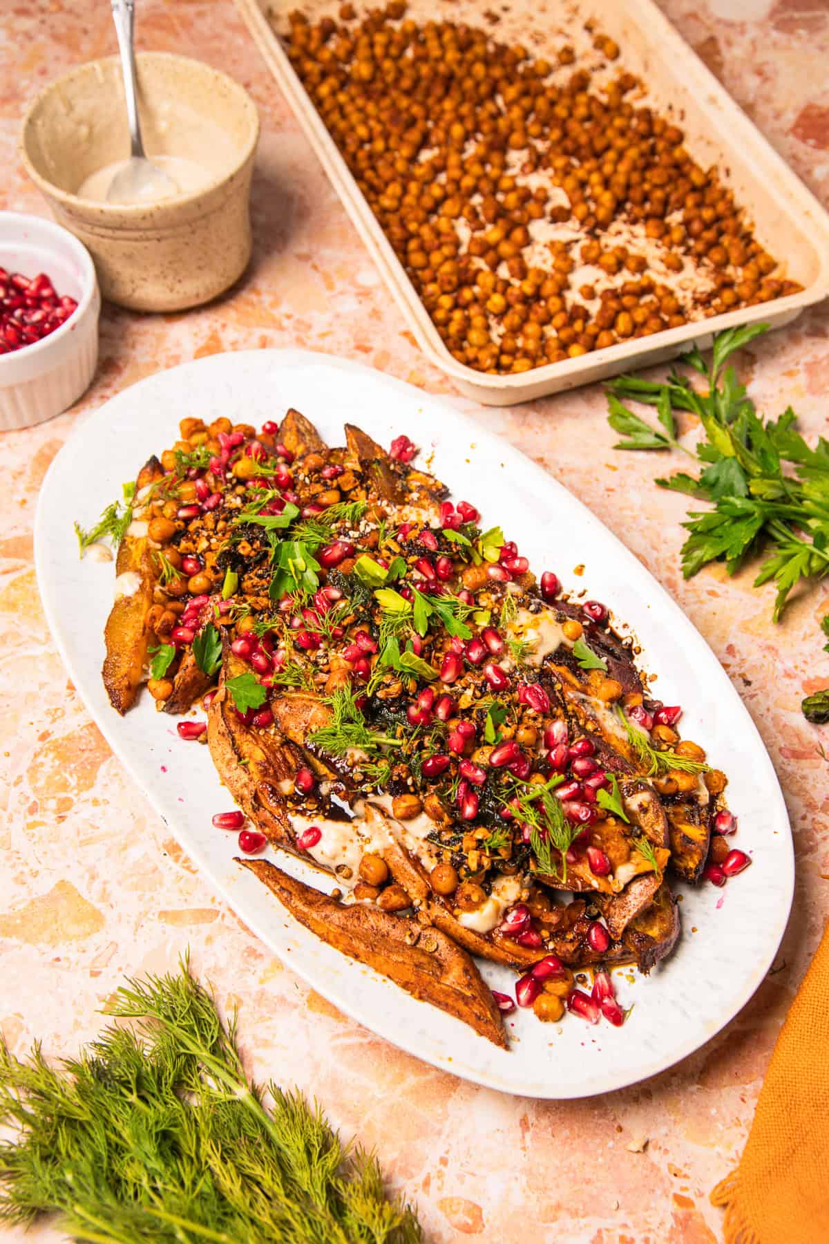 Roasted eggplant topped with chickpeas, pomegranate seeds, green herbs, and sesame seeds on a white plate—perfect alongside roasted sweet potatoes with tahini.