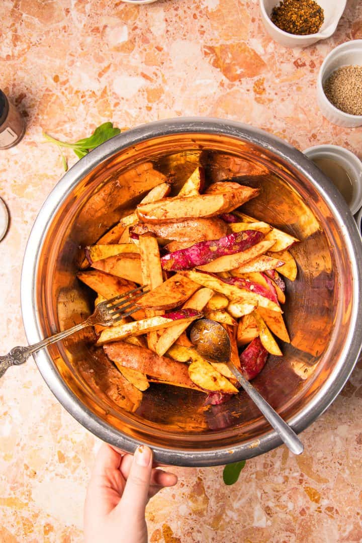 Hand mixing seasoned potato wedges in a metal bowl with two spoons on a marble countertop, perfect for creating roasted sweet potatoes with tahini.