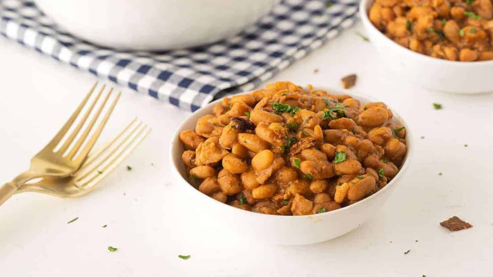 A bowl of baked beans garnished with herbs sits next to gold forks and a checkered napkin.