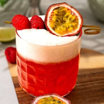 A red cocktail with foam, garnished with raspberries and half a passion fruit, on a wooden tray with lime slices.
