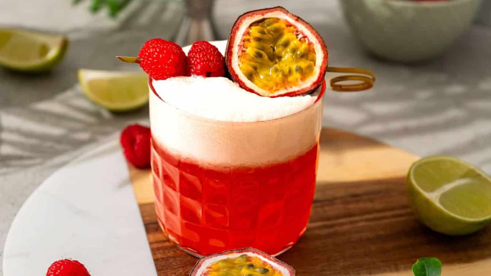 A red cocktail with foam, garnished with raspberries and half a passion fruit, on a wooden tray with lime slices.