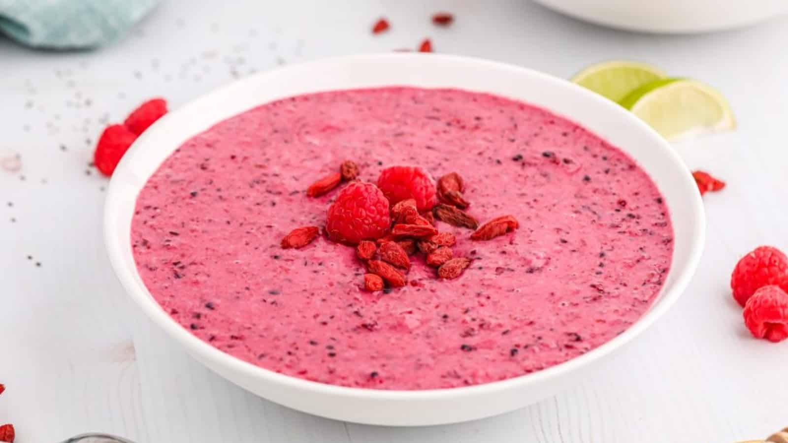 A white bowl filled with a pink smoothie, topped with raspberries and goji berries.
