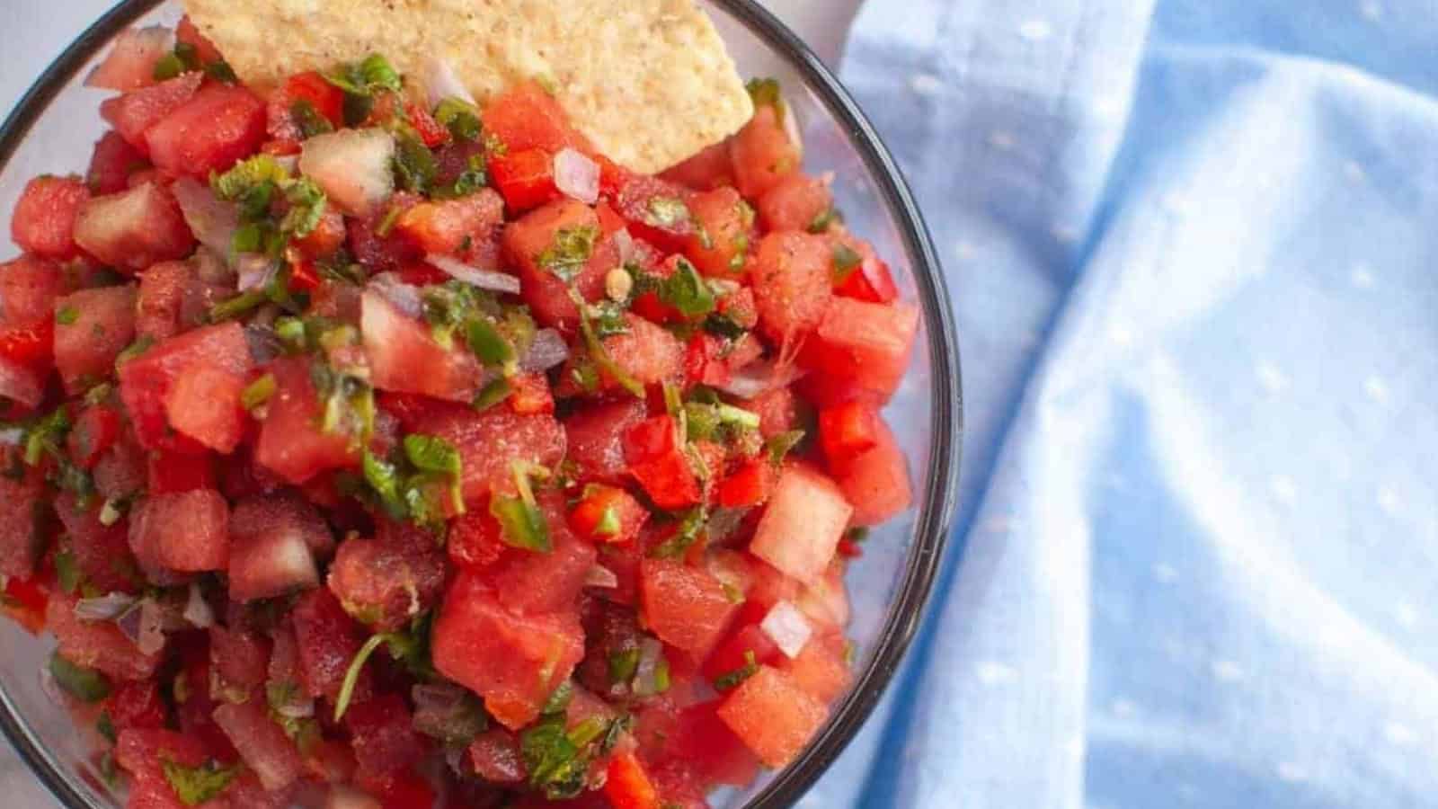 A bowl of watermelon salsa with chopped herbs and vegetables, served with a tortilla chip, next to a light blue cloth.