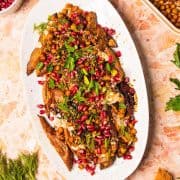 Roasted eggplant on a white platter topped with chickpeas, herbs, pomegranate seeds, and sauce—perfect alongside roasted sweet potatoes with tahini for a vibrant, flavorful spread.
