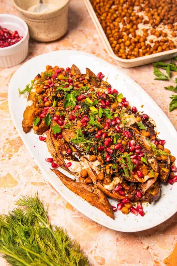 White platter with roasted sweet potatoes with tahini, chickpeas, herbs, and pomegranate seeds on a marble surface.
